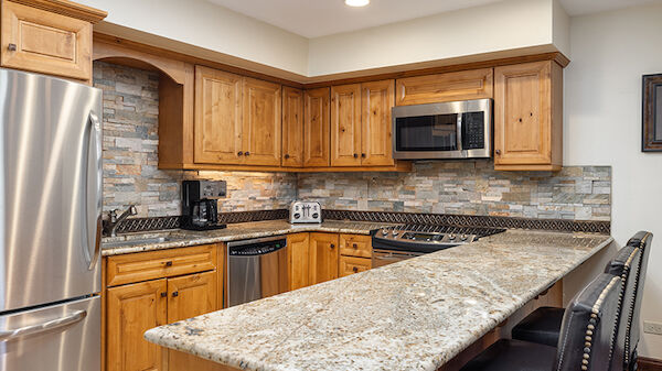 A kitchen with wooden cabinets, stainless steel appliances, and a long granite countertop with stools.