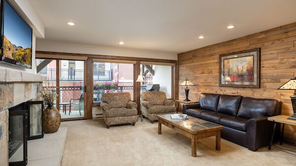 Cozy living room with stone fireplace, beige carpet, tan chairs, a dark leather sofa, wooden walls, coffee table, and warm lamps, plus a balcony view.