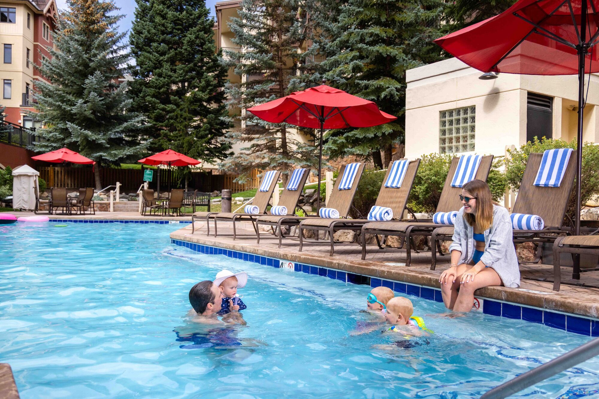 A resort pool scene: a mother and toddler swim with floating devices, sun loungers, red umbrellas, and striped chairs by the water.