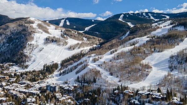 The image shows a snowy mountain landscape with ski slopes, a nearby town, and scattered trees under a blue sky with clouds.