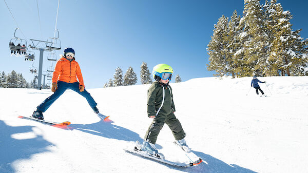 People skiing downhill on a snowy slope with a chairlift in the background; child in green pants leads, wearing a yellow helmet, orange jacket nearby, sunny day.