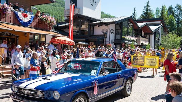 A bright blue vintage car drives through a busy outdoor crowd with spectators, banners, and wooden buildings in a small town setting.