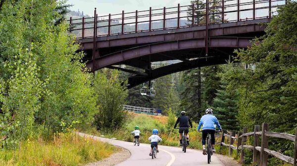 Riverside bike path under a wooden bridge, people biking together on a scenic forest trail.