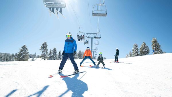 Snowy ski slope with skiers, a chairlift in the background, clear blue sky, and sun glare; trees line the horizon as shadows stretch long.