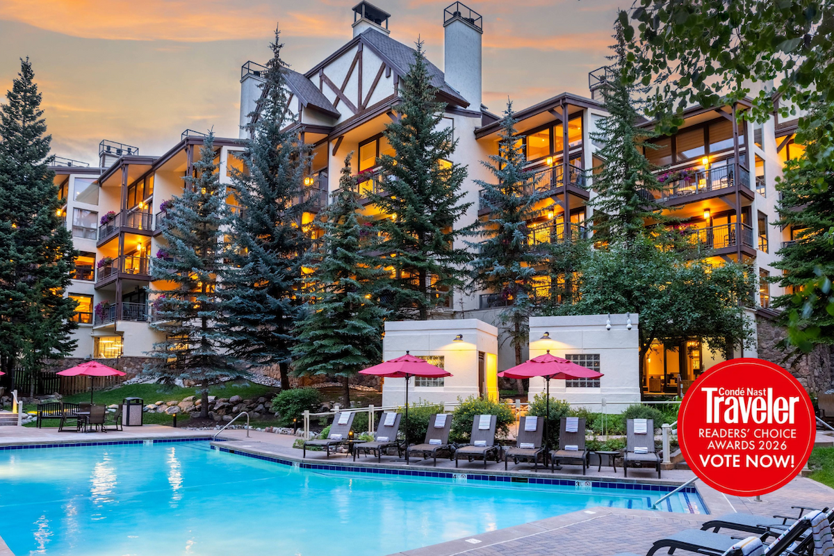 A resort-style hotel with a bright pool, lounge chairs, red umbrellas, tall evergreen trees, and warm evening lighting. End.