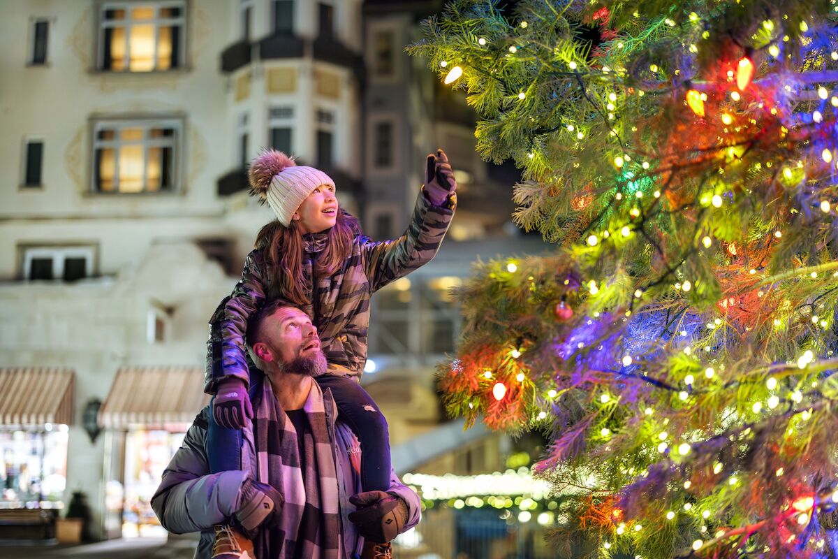 A couple outdoors at night, man carrying a child on shoulders, admiring a brightly lit Christmas tree with colorful lights. End.