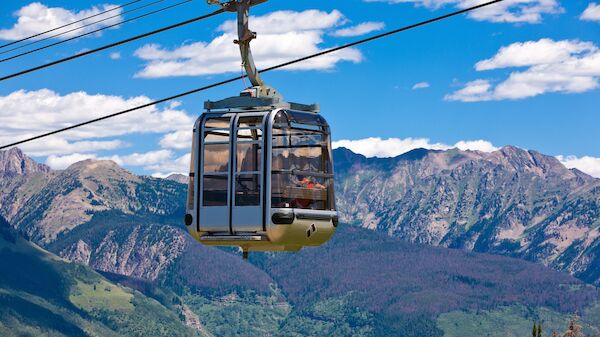 A cable car glides over a lush green valley with rugged mountains under a bright blue sky, hanging from cables high above the forested slopes.