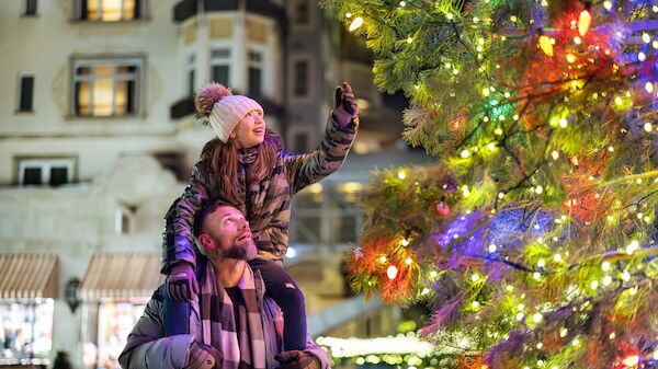 A couple in a city square admire a brightly lit Christmas tree; the man carries the child on his shoulders as lights glow around them.