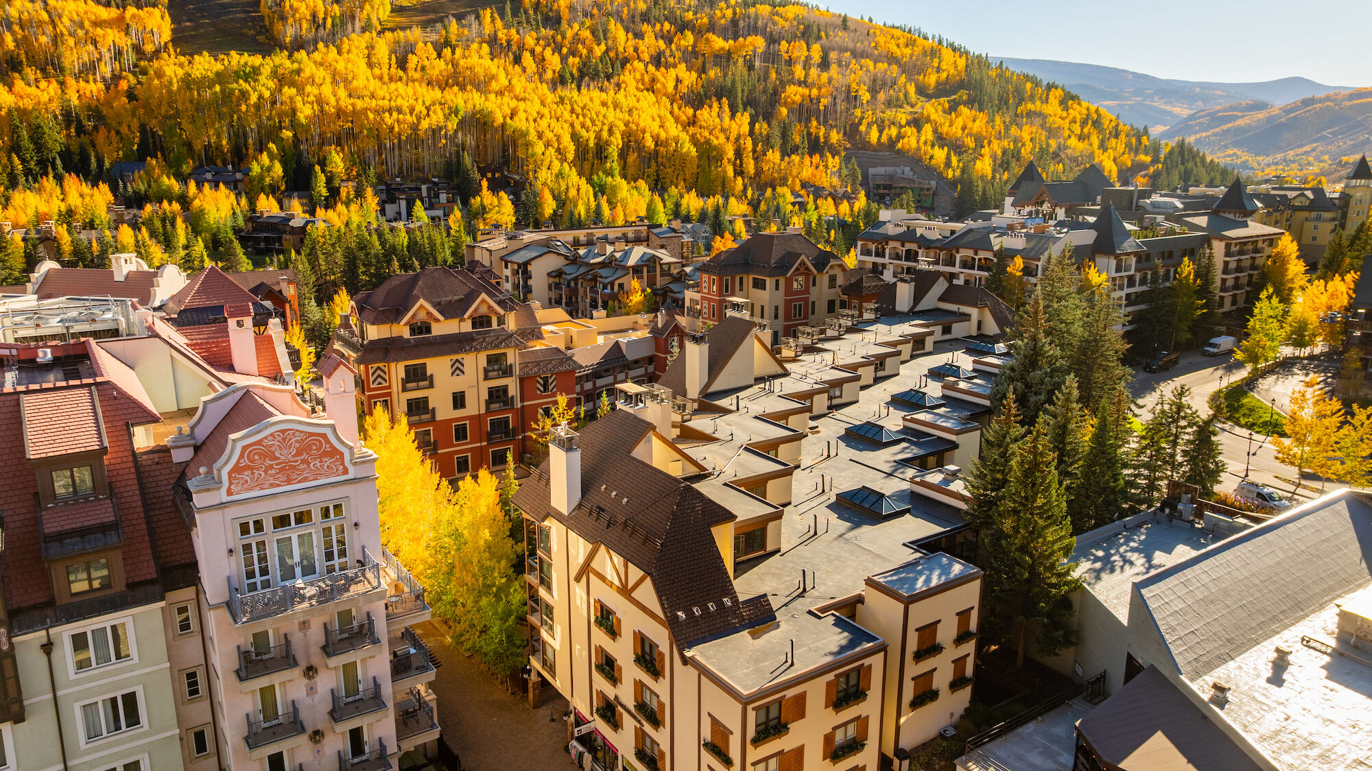 Aerial view of a mountain town with colorful autumn trees, clustered chalet-style buildings, and winding streets under golden foliage.
